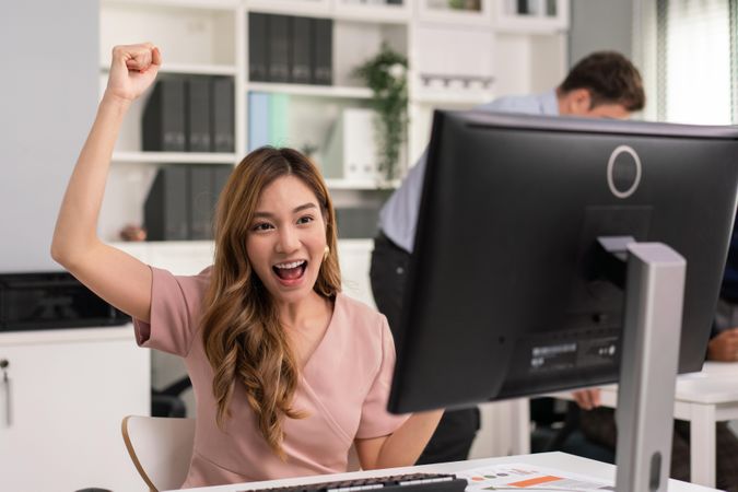 Asian woman with arm up in celebration while sitting at her desk