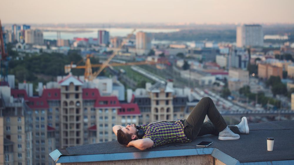 Male lying back looking at sky, with tablet, headphones and takeaway coffee cup on roof