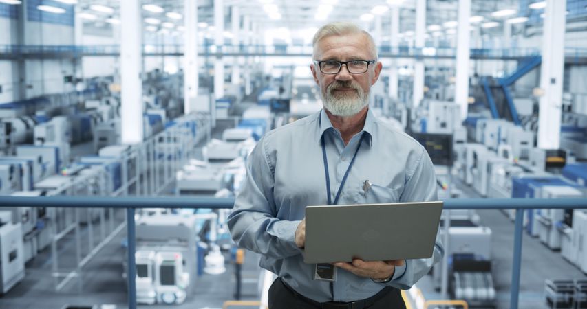 Portrait of a Mature Technician with a Beard and Glasses Using Laptop Computer and Looking at Camera. Industrial Male Specialist Wearing a Protective Vest, Working in a Modern Electronics Factory