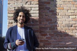 Smiling Black man holding his smartphone while leaning on a brick wall outdoors on sunny day 0PjG37