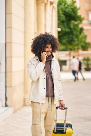 Portrait of a young man with afro hair talking on the phone on the street with yellow suitcase
