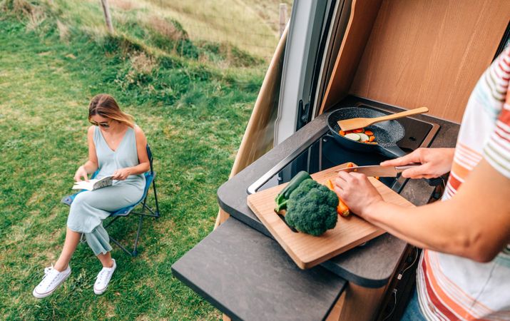 Male chopping vegetables in motorhome kitchen with door open to woman reading outside