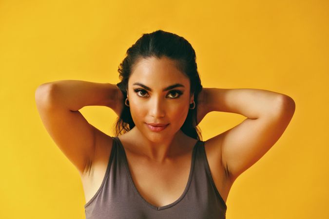 Headshot of Hispanic woman looking at camera with both hands at back of head on yellow background