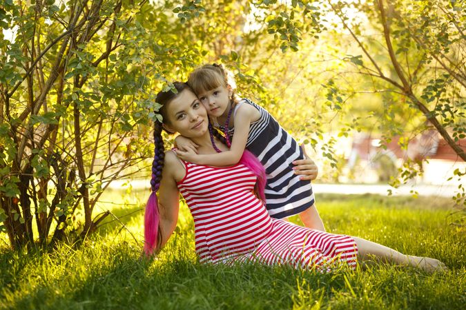 Pregnant female in red striped dress holding her daughter under the trees while sitting on grass