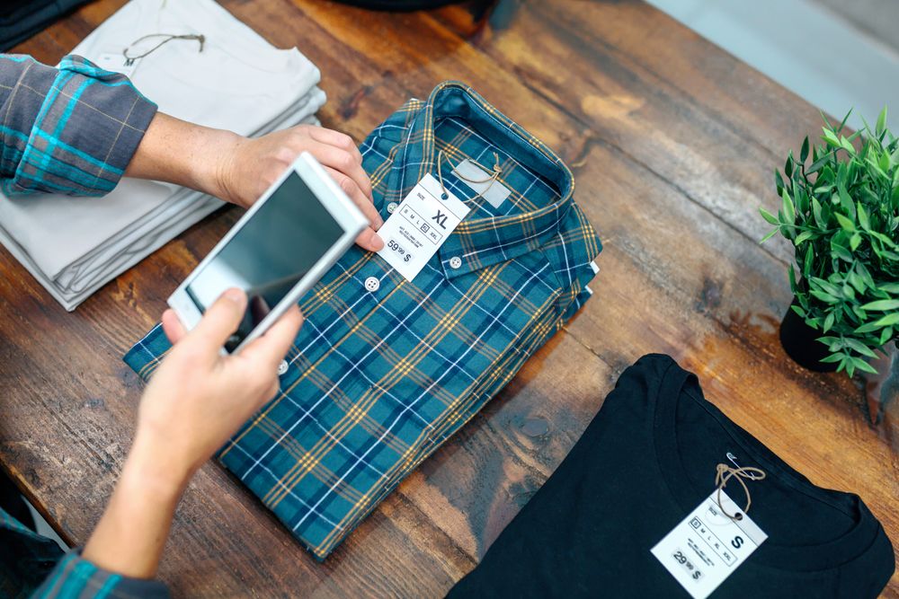 Unrecognizable woman customer holding cellphone while scan label of plaid shirt in clothes shop