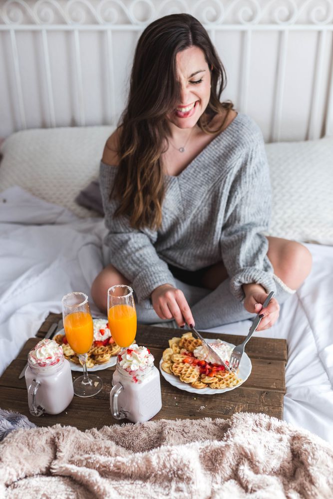 Young woman eating appetizing waffles and tasty drinks on tray on bed