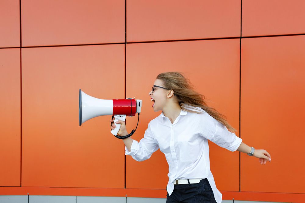 Woman in glasses in front of orange wall speaking into megaphone