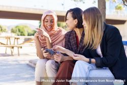 Three happy women sitting on outdoor park bench talking while watching smartphone 4NgAeb