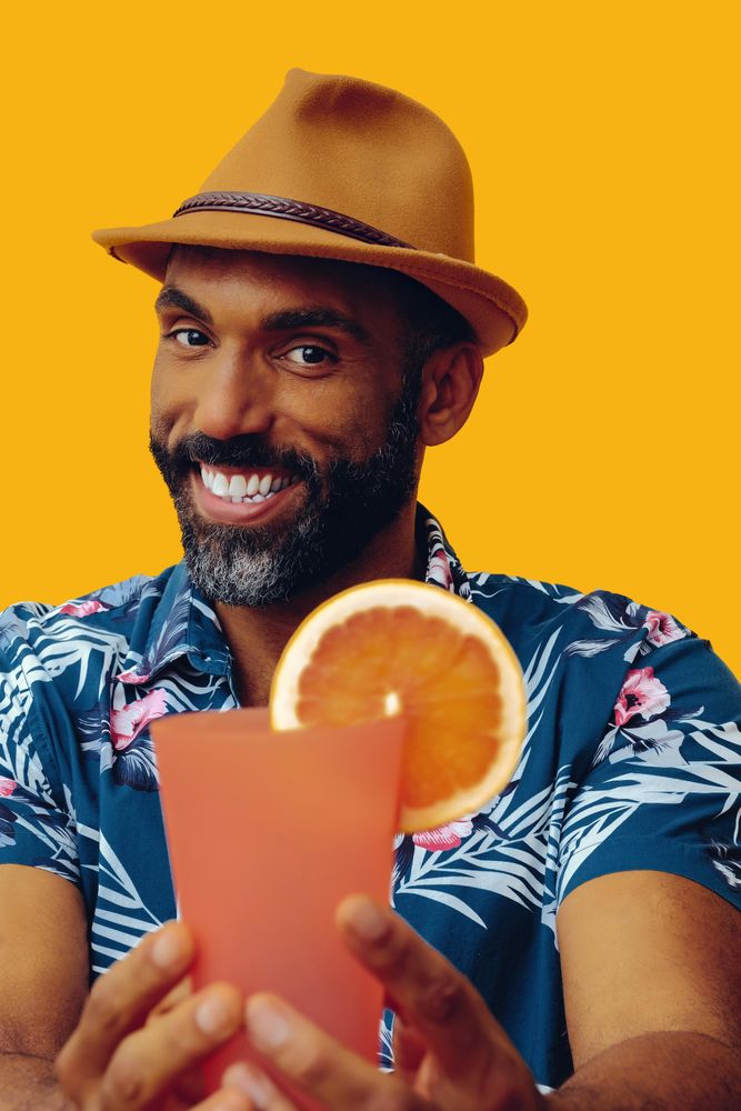 Male passing a cocktail to camera while wearing colorful shirt with yellow background