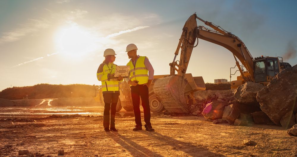 Hispanic Female Urban Planner Using Laptop And Talking To Male Real Estate Investor On Construction Site. Colleagues Discussing New Project While Heavy Machinery Working. Warm Edit