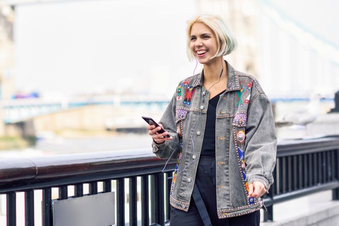 Cheerful blonde woman listening to music on phone along river walk