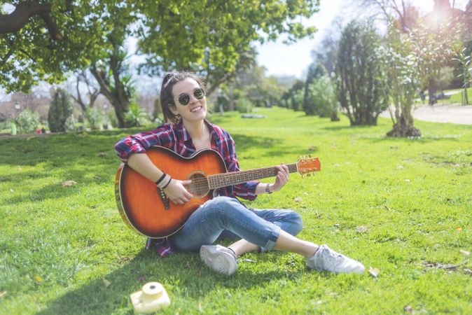 Front view of a young hipster woman wearing sunglasses, sitting on grass in a park while enjoying playing guitar