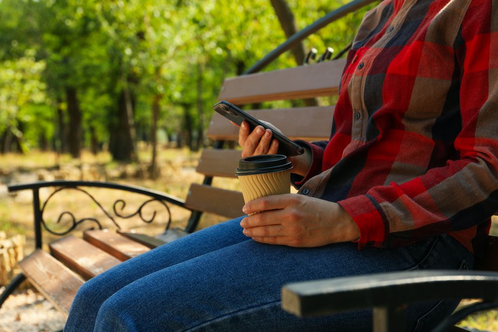 Woman in jeans and shirt hold paper cup and phone outdoor