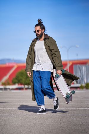 Trendy hipster man strolling on asphalt road with skateboard