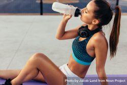 Female sitting on exercise mat and drinking water while exercising 0LLaD0
