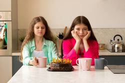 Two girls sitting with birthday cake and mugs at kitchen.