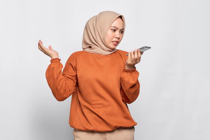 Worried Muslim woman talking on smart phone speaker and making gesture with hand up