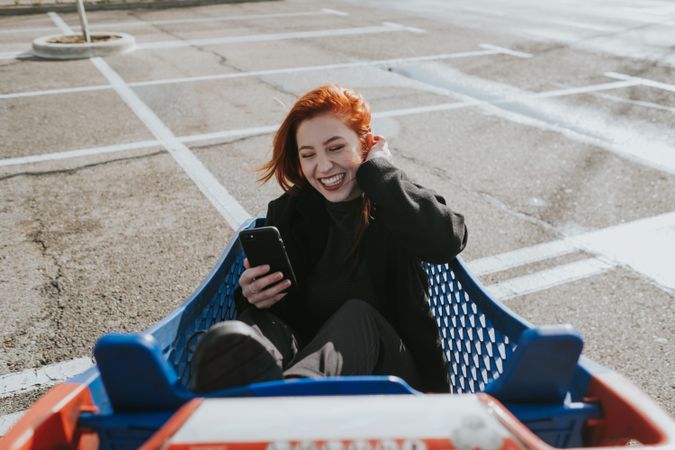 Smiling woman with smartphone in shopping trolley in parking lot