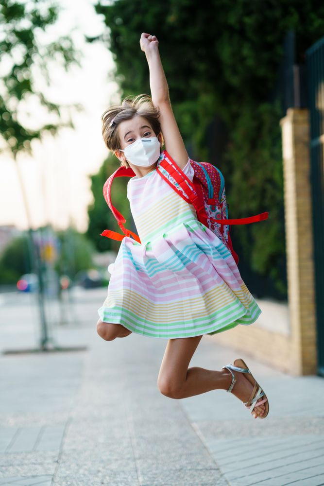 Happy girl jumping outside school gates wearing facemask