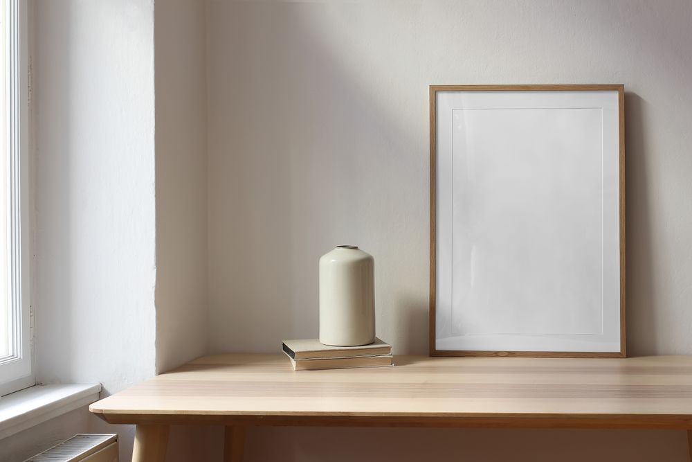 Living room with ceramic vase, old books on wooden table near window, with blank wooden picture frame mockup