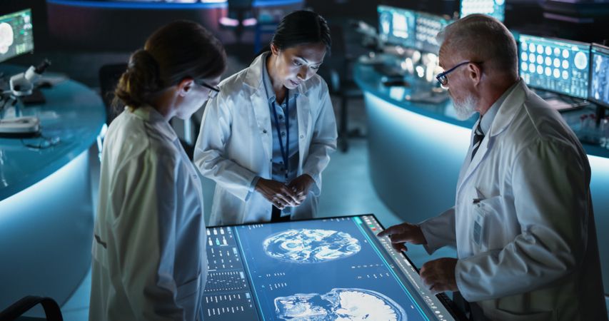 Modern Hospital Medical Research Center: Diverse Colleagues Gathered Around Interactive Touch Screen Table With MRI Scans Of Brain On Display.