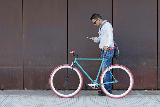 Male in sunglasses standing with red and green bicycle looking down at cell phone