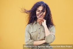 Female in army green jumpsuit smiling in front of mustard wall with hair blowing 0W7z60