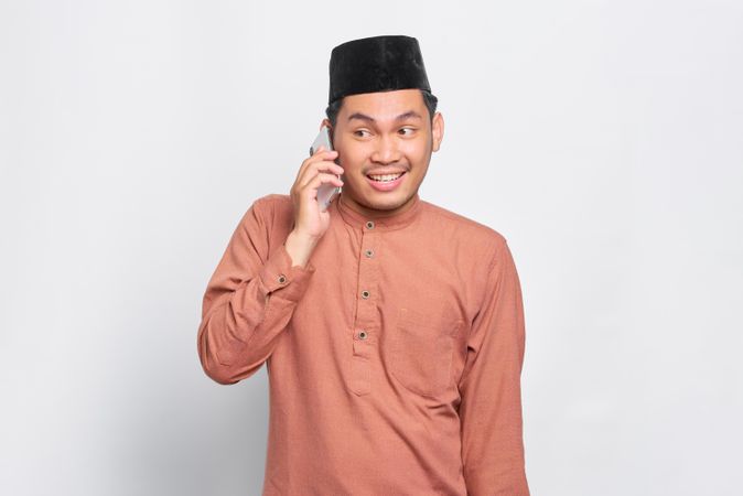 Excited Muslim man in kufi hat smiling and talking on smart phone