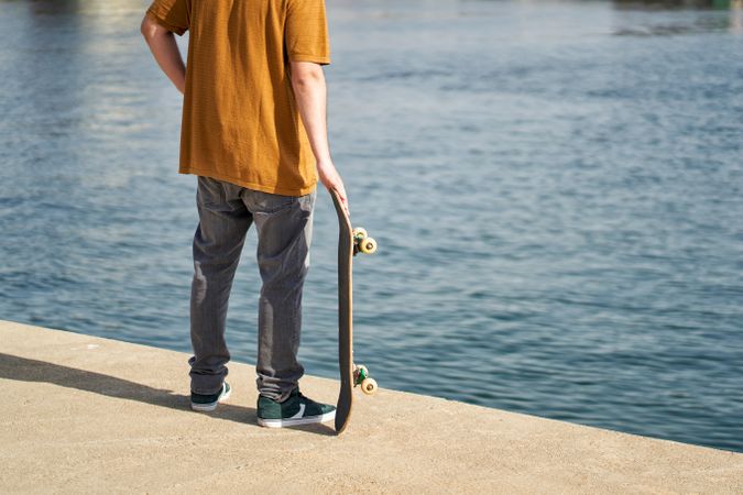an unrecognizable person standing with his skateboard in his hand looking out to sea