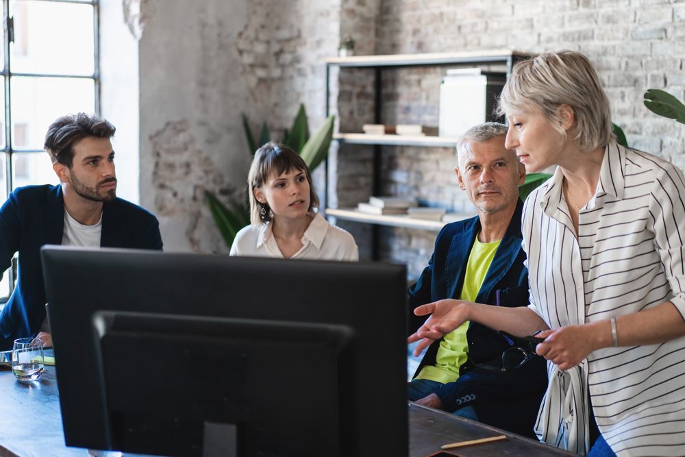 Co-workers sitting around a desk in a modern office, discussing a project and looking at a computer monitor