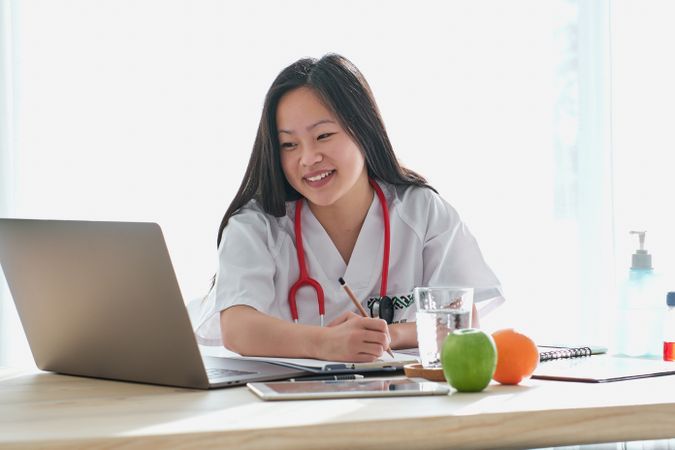 Cheerful Asian doctor browsing laptop in medical office