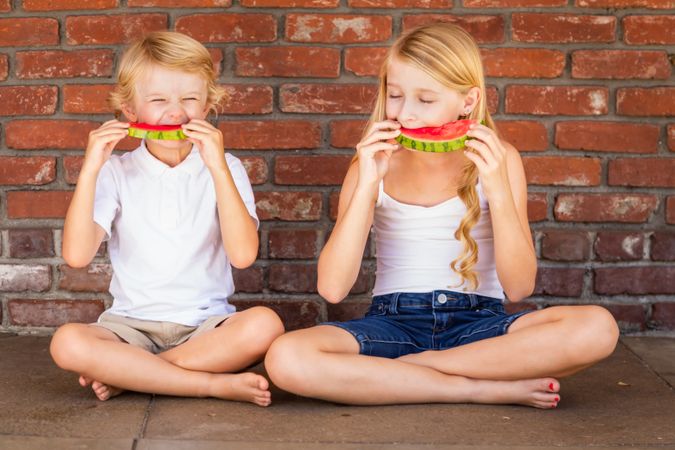 Cute Young Boy and Girl Eating Watermelon Against Brick Wall