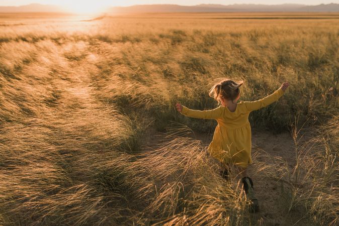 Young girl in yellow dress walking in tall grass field at sunset