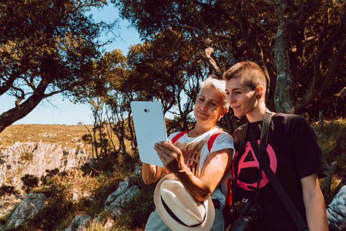 Older woman and punk woman looking at tablet sitting on stone in valley