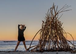 Young woman dancing on shore near water and wooden twigs placed for campfire