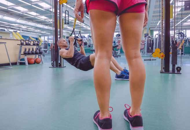 Legs of female trainer looking on at person using calisthenics in gym