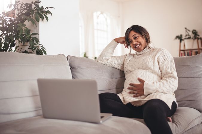 Woman expecting baby sitting on couch using laptop at home