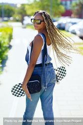 Female in denim overalls standing with skateboard and looking behind her beZq60
