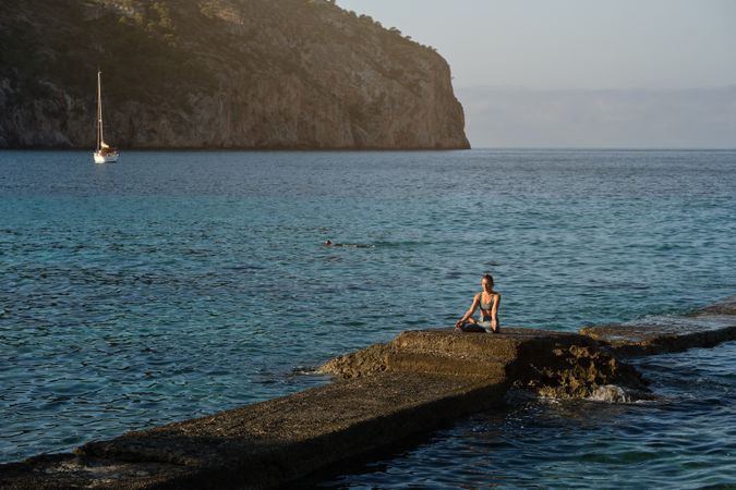 Woman sitting on boulder among blue ocean and taking Padmasana p