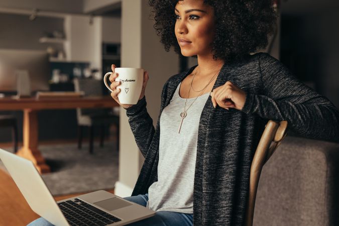 Young woman sitting in living room with laptop and cup of coffee looking away thinking