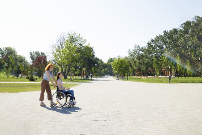 Red haired woman pushing her friend’s wheelchair in a public park