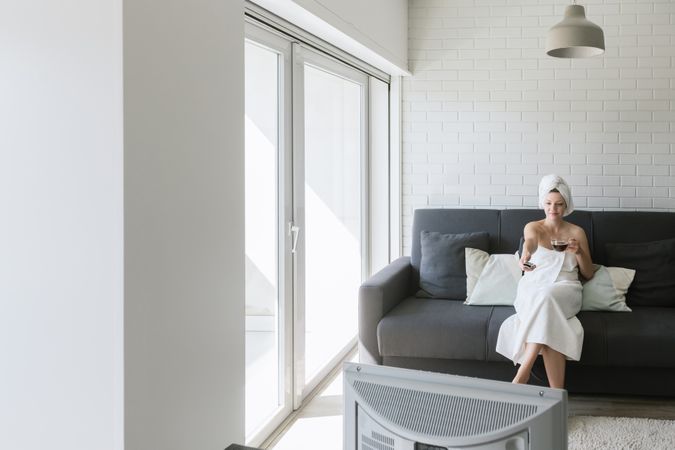 Happy relaxed woman enjoying day off while having hot drink after shower and watching TV in apartment in loft style