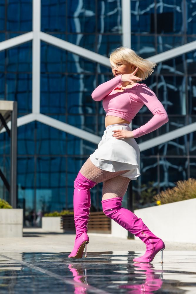 Stylish Woman In Pink Top And White Skirt Paired With Pink Thigh-High Boots Strikes A Pose Against A Modern Glass Building Background.