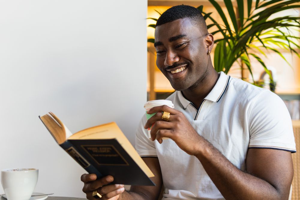 Smiling Black man reading book with drink in cafe