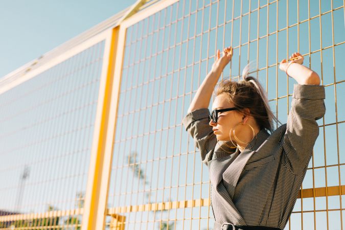 Stylish woman in sunglasses raising hands and leaning on lattice