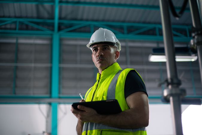 An industrial engineer wearing a safety helmet and reflective vest is standing inside a factory while holding a digital tablet. He appears to be monitoring operations or conducting an inspection