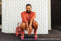 Excited man in makeup smiling at the camera cheerfully while squatting against a brick background 48DrJ4
