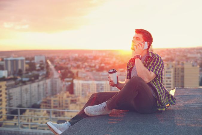 Male taking phone call while sitting on roof with takeaway coffee