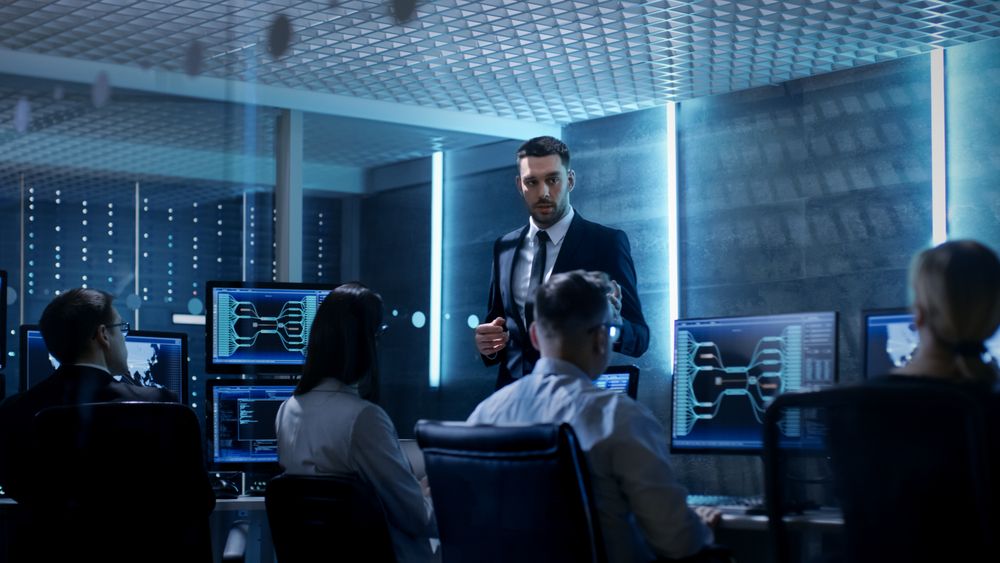 Supervisor Holds Briefing for His Employees in System Control Center Full of Monitors and Servers.