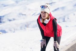 Focused fit woman in warm clothes and headphones leaning on knees while wintry road